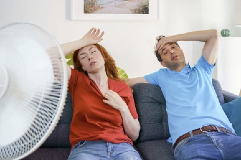 A decorative photo of a couple sitting on their sofa trying to cool down in front of a portable fan. They need a whole house fan!