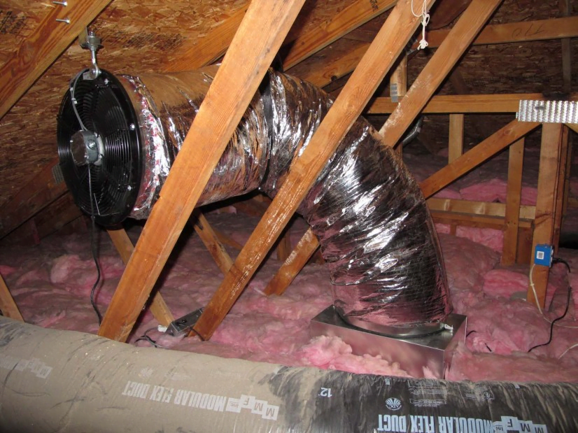 A photo of a CentricAir ducted whole house fan installed in an attic, showing how the fan is attached to the attic framing, and that the fan itself is at the end of a duct and around a corner to so that it is very quiet.